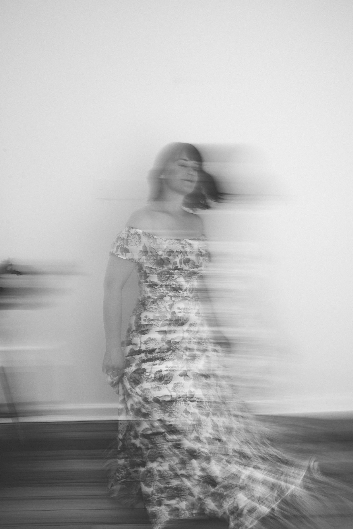 A black and white motion blur image of a lady in a full length floral dress- she appears to be moving backwards.