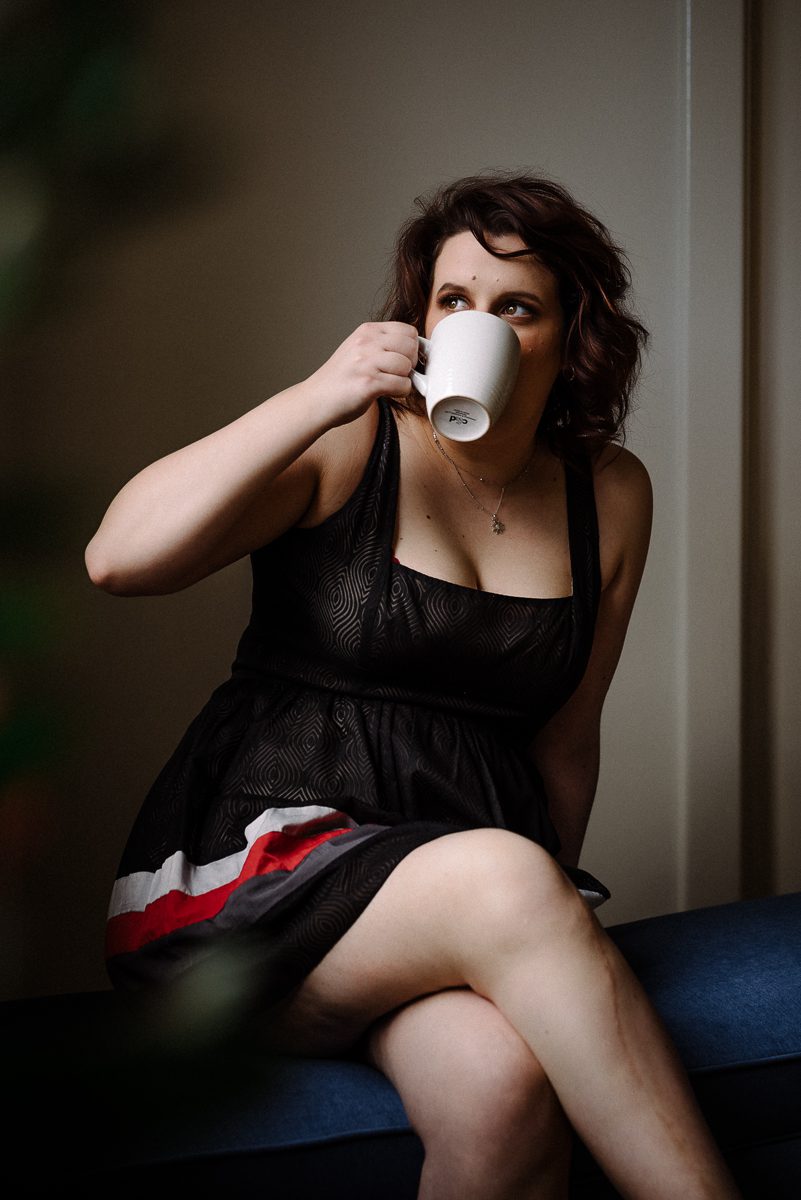 A photograph of a woman in a black dress with coloured stripes at the bottom. She is sitting and sipping from a white tea cup while she looks out the window.