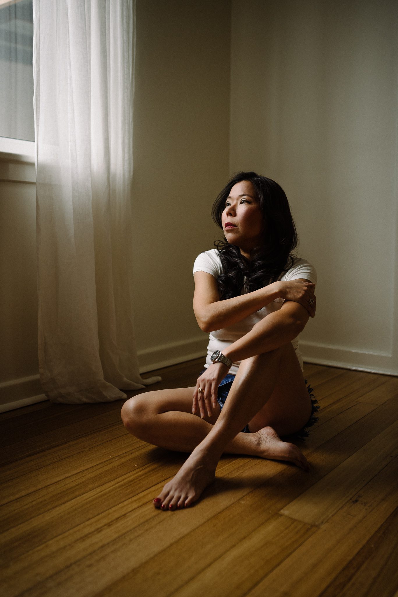 A lady sitting on the floor with her arms crossed, the light is hitting her face and the side of her body - taken during a boudoir shoot.